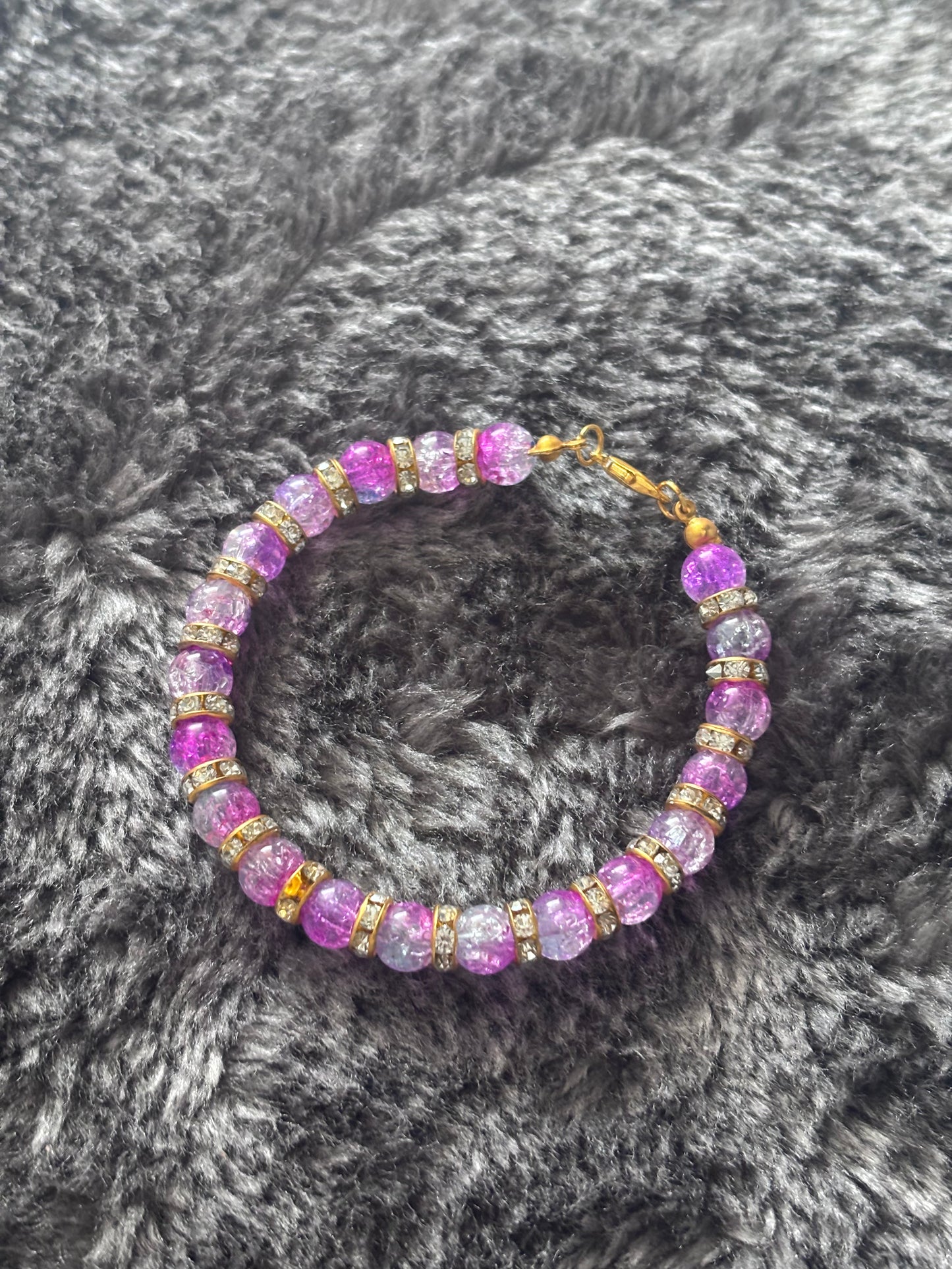 Handgemaakte armband met paarse gebarsten glaskralen en goudkleurige strass ringen, liggend op een zachte grijze ondergrond.