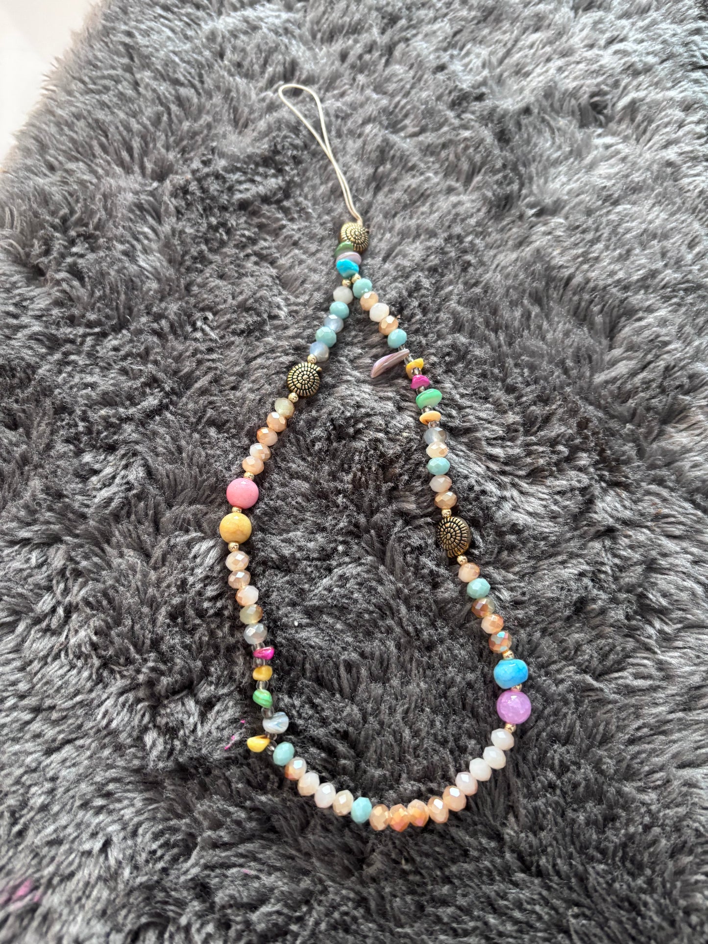 Handgemaakt kleurrijk telefoonkoord met kralen in pastel- en aardetinten op een grijze zachte achtergrond.