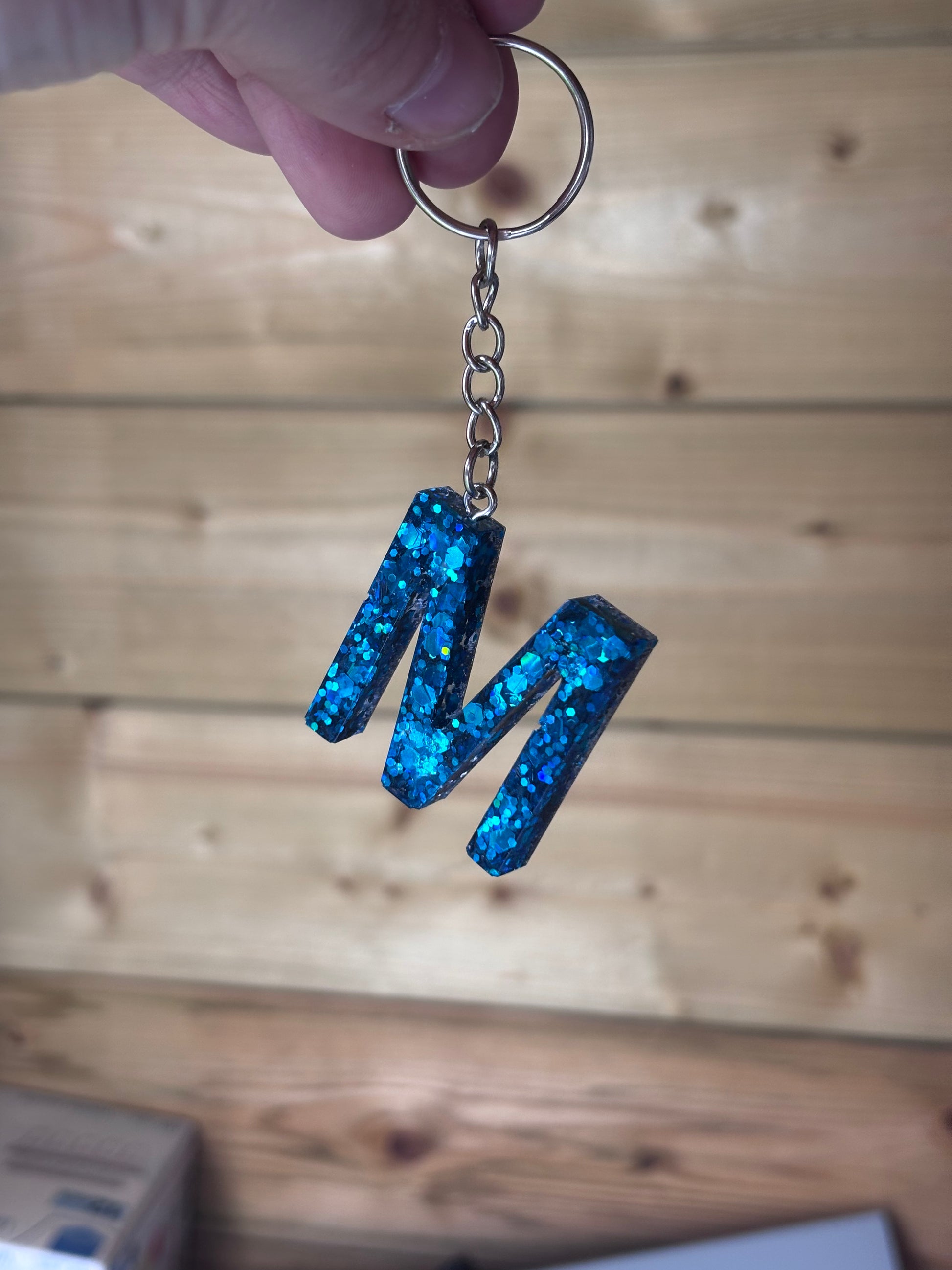Handgemaakte epoxy sleutelhanger in de vorm van de letter “M”, gevuld met sprankelende blauwe glitter, vastgehouden tegen een houten achtergrond.