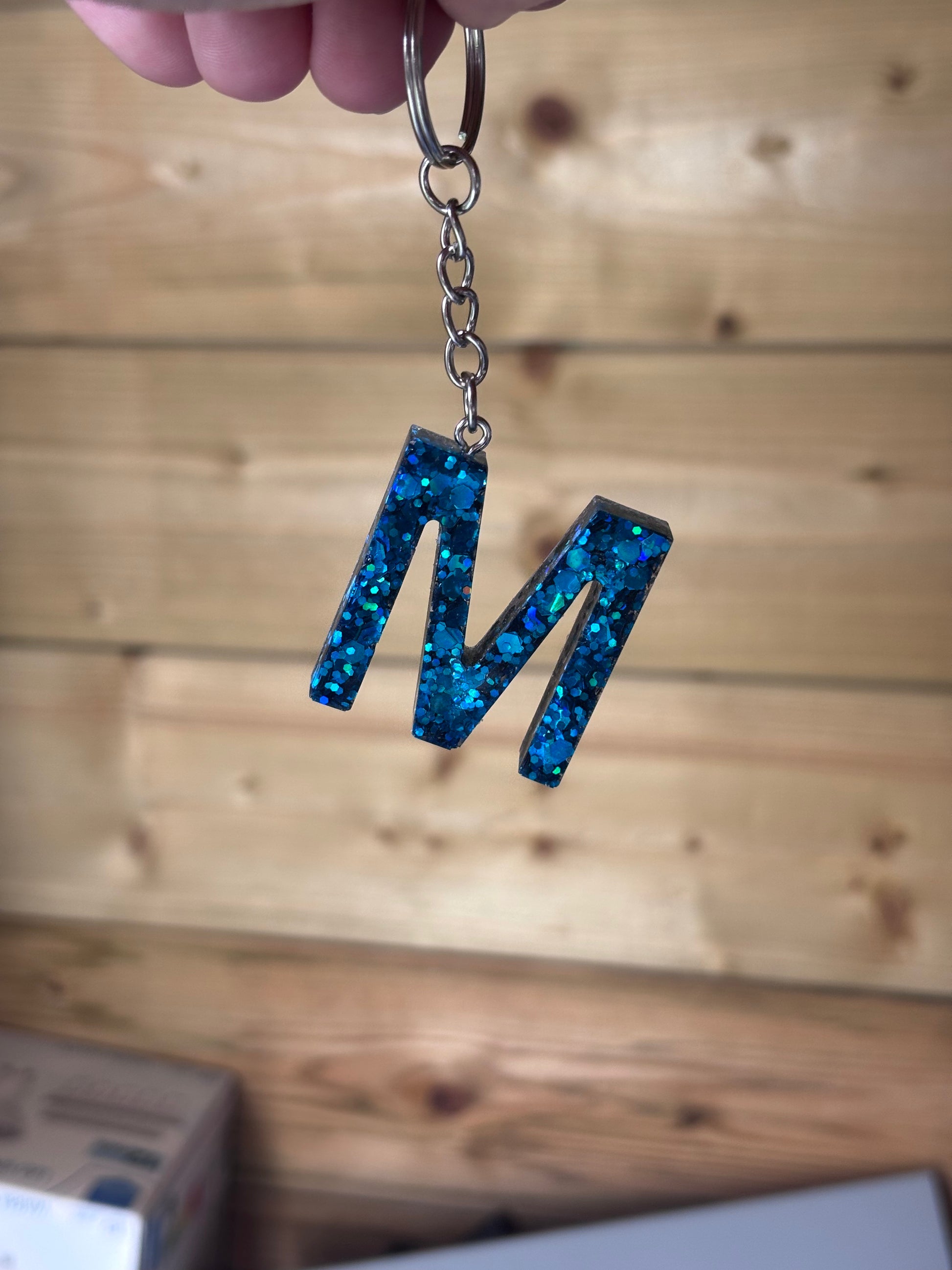Handgemaakte epoxy sleutelhanger in de vorm van de letter “M”, gevuld met sprankelende blauwe glitter, vastgehouden tegen een houten achtergrond.