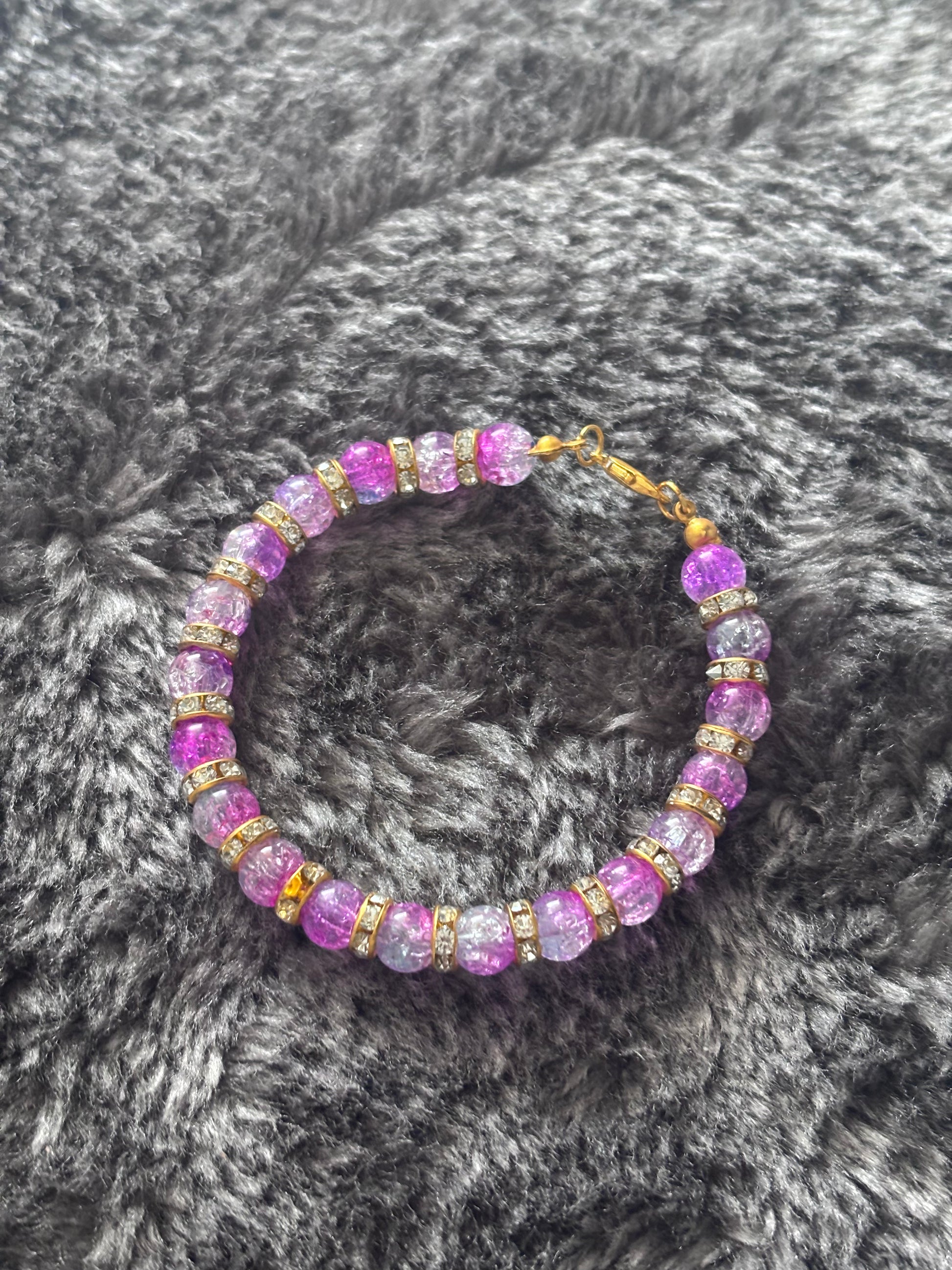 Handgemaakte armband met paarse gebarsten glaskralen en goudkleurige strass ringen, liggend op een zachte grijze ondergrond.