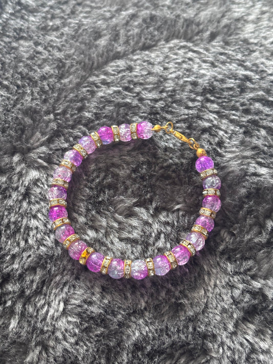 Handgemaakte armband met paarse gebarsten glaskralen en goudkleurige strass ringen, liggend op een zachte grijze ondergrond.