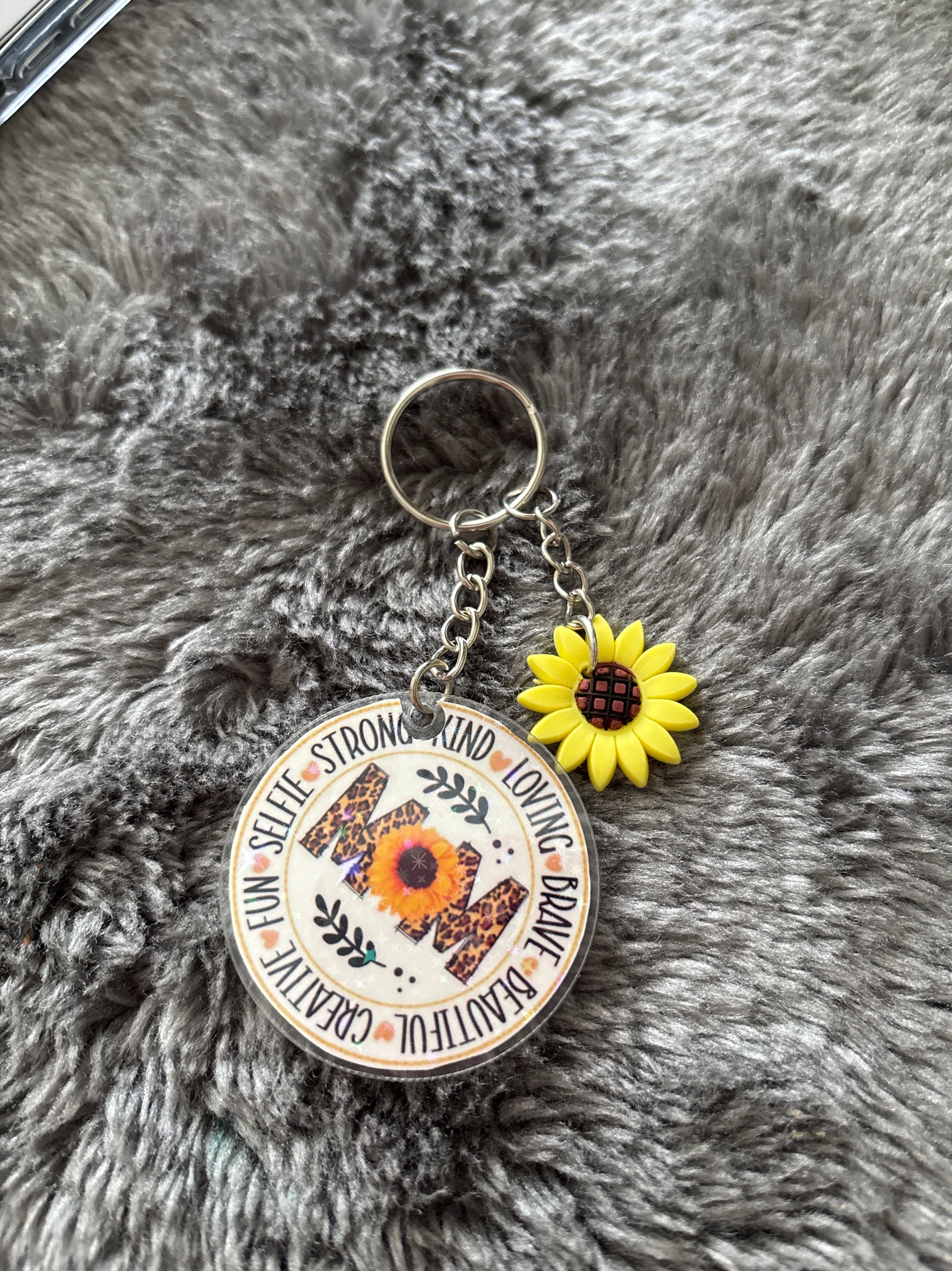 Sleutelhanger met het woord “MOM” versierd met een zonnebloem en luipaardprint, omringd door inspirerende woorden, met een extra kleine zonnebloembedel.