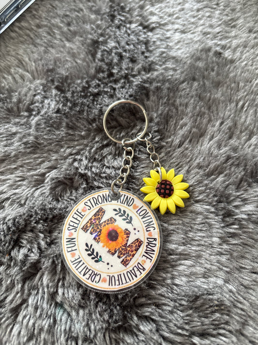 Sleutelhanger met het woord “MOM” versierd met een zonnebloem en luipaardprint, omringd door inspirerende woorden, met een extra kleine zonnebloembedel.